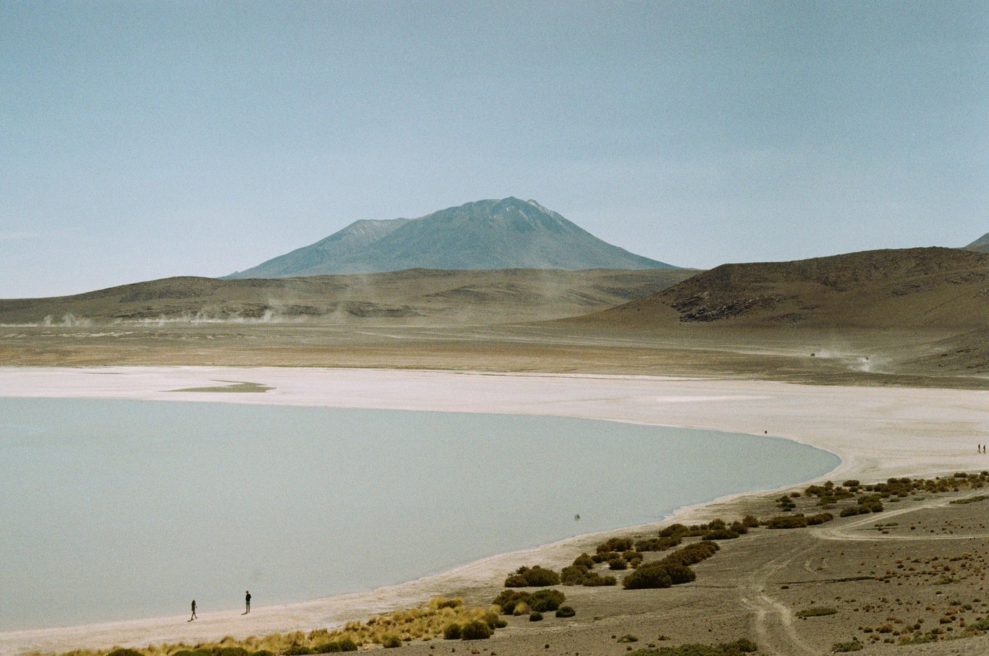 Two people stand on the shore of a pale lake with a large mountain in the background.