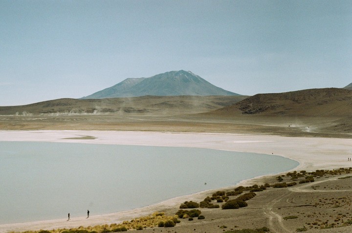 Two people stand on the shore of a pale lake with a large mountain in the background.