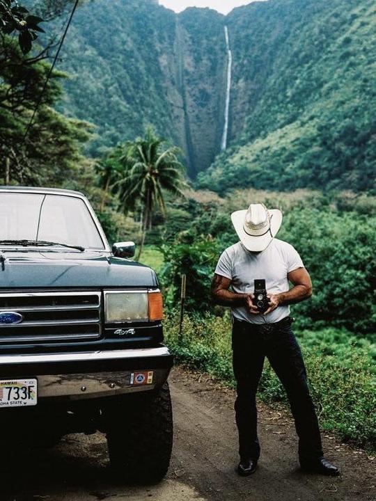 A man in a cowboy hat stands next to a truck, holding a camera, with a waterfall and lush mountains in the background.
