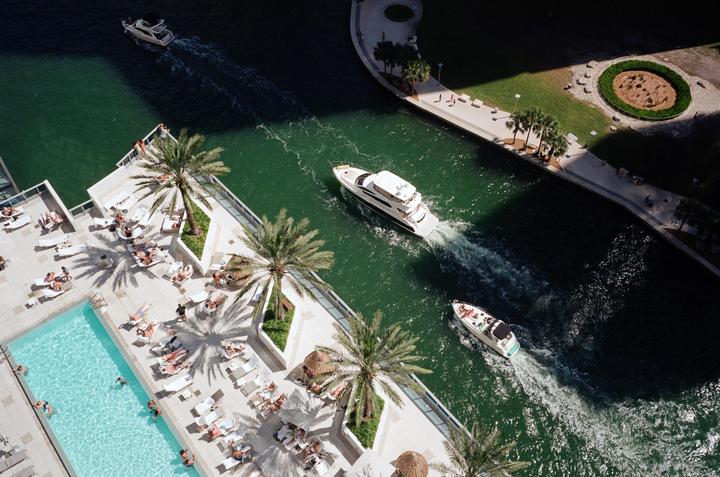 An aerial view shows boats on a canal next to a building with a swimming pool and people sunbathing.