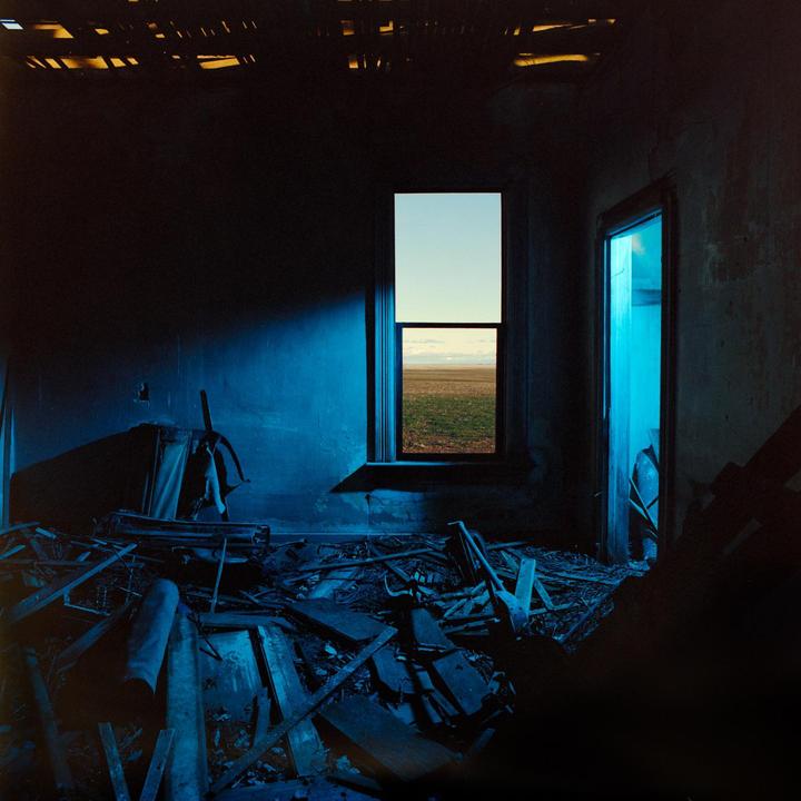 A dark, ruined room with blue light contains a window looking out onto a field.