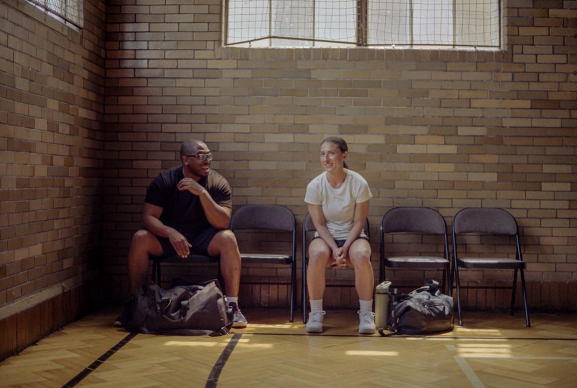 A man and a woman sit on chairs in a room with a brick wall and a wooden floor.