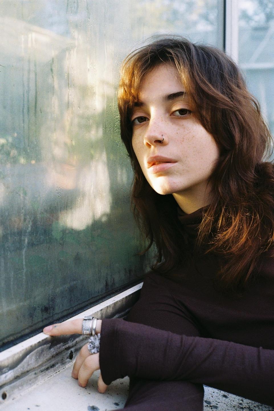 A woman with long hair and freckles looks at the viewer while leaning against a wet window.