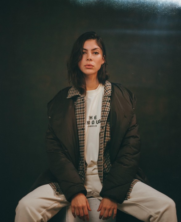 A woman with dark hair sits wearing a plaid shirt, a dark jacket, and light-colored pants.