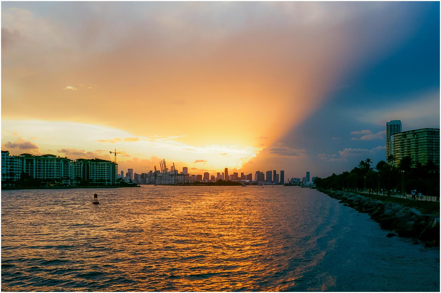 A vibrant sunset over the Miami skyline casts a dramatic ray of light across the water.