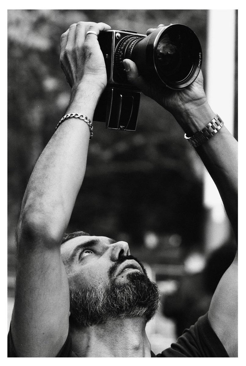 A man with a beard holds a camera above his head, looking up at it.