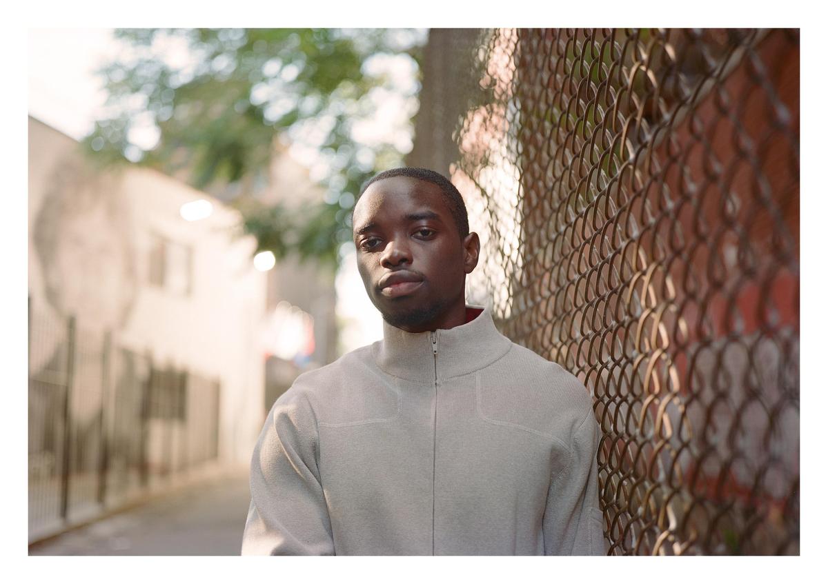 A man in a light-colored jacket stands next to a chain-link fence in an alley.