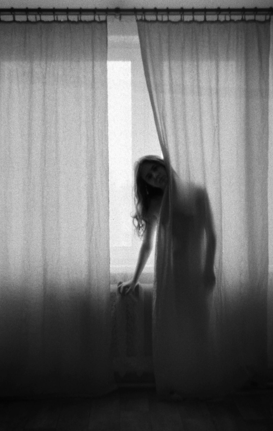 A woman stands behind a sheer curtain, looking out a window.