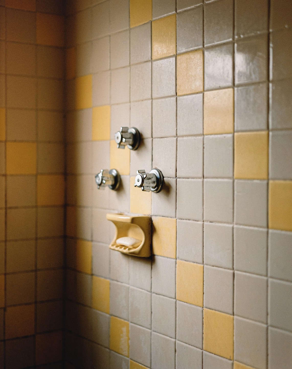 A yellow soap dish with a bar of soap is mounted on a tiled wall with three metal knobs.