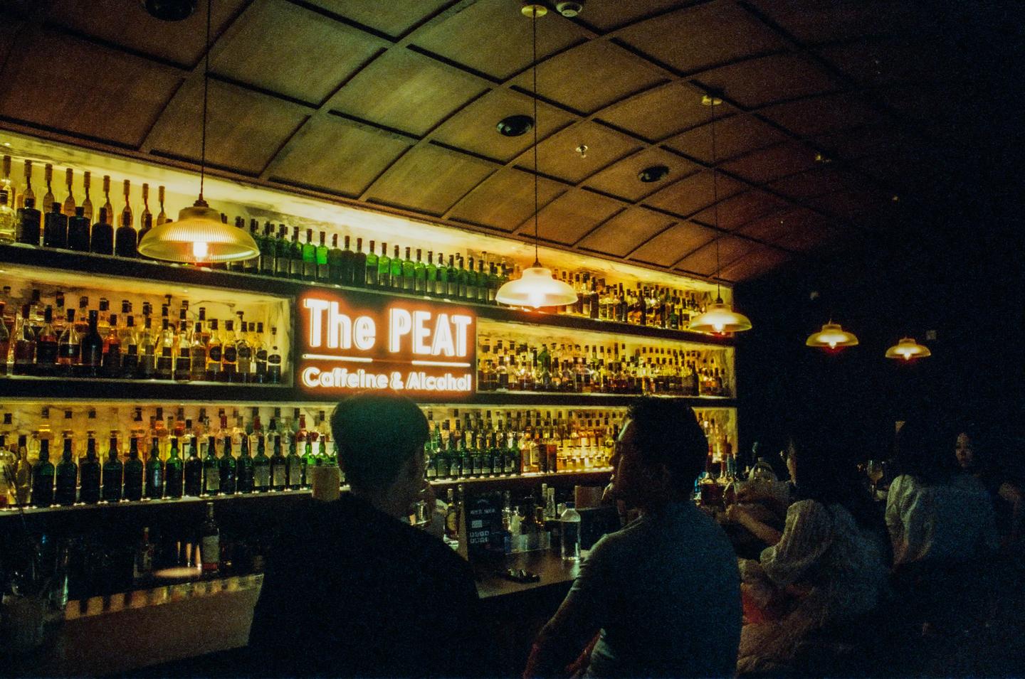 People sit at a bar with a large display of liquor bottles and a neon sign that reads "The PEAT Caffeine & Alcohol."