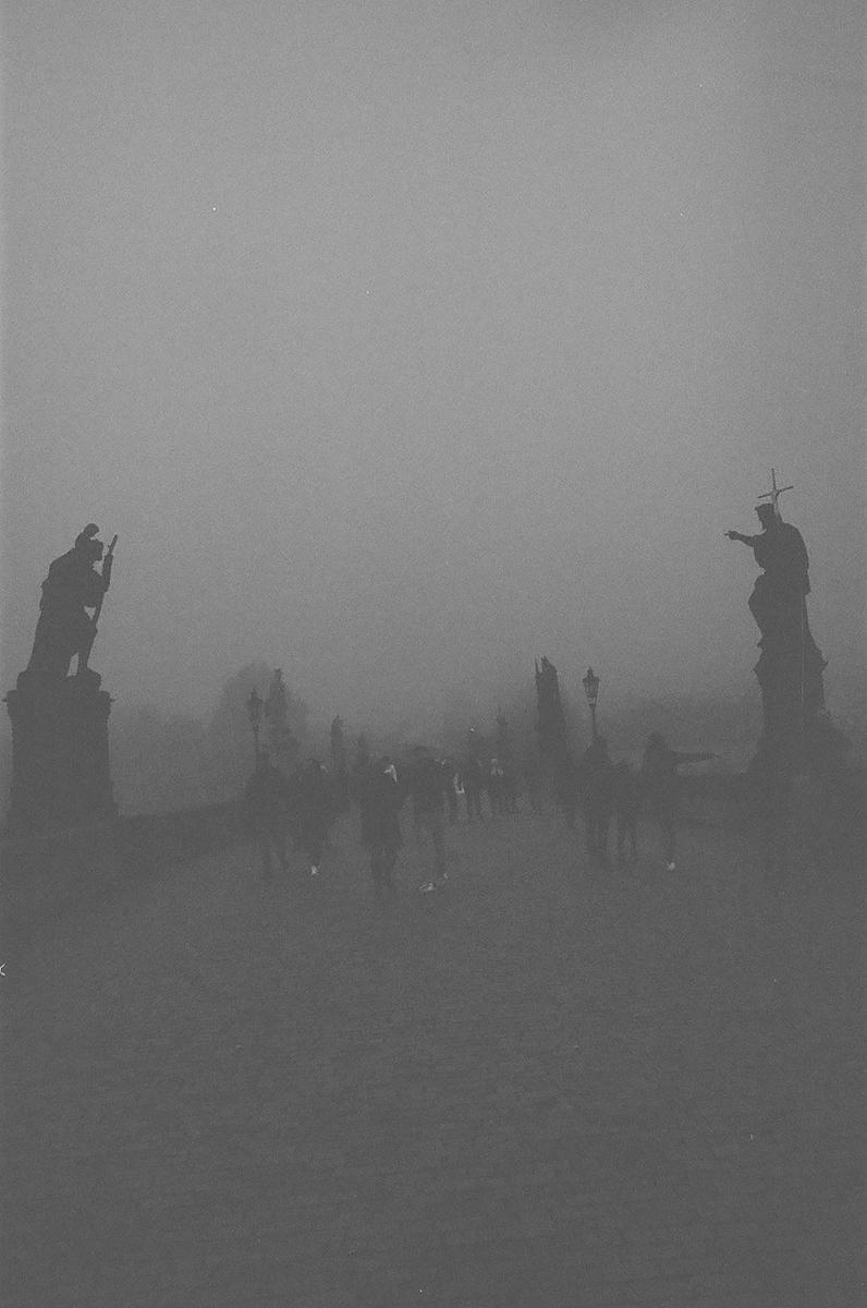 People walk on a bridge with statues on a foggy day.