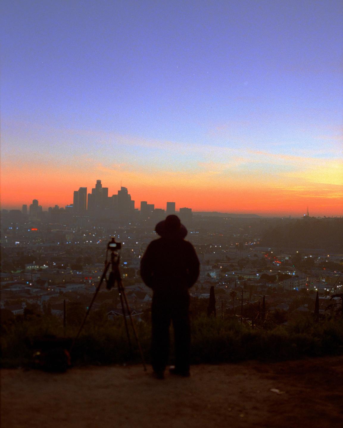 A person stands on a hill overlooking the Los Angeles skyline at sunset, with a tripod set up nearby.