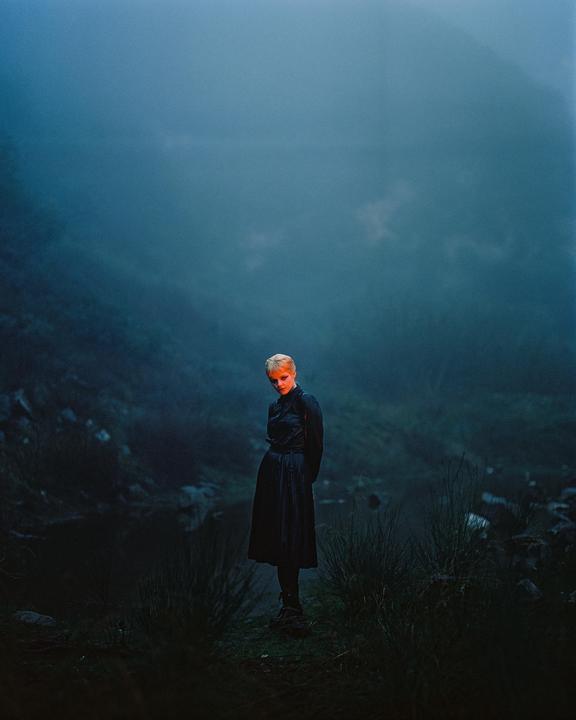 A woman with short blonde hair stands in a dark, misty landscape with a red glow on her face.
