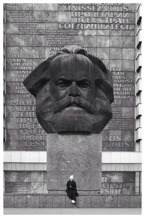 A woman sits on a ledge in front of a large statue of Karl Marx, with text carved into the wall behind it.