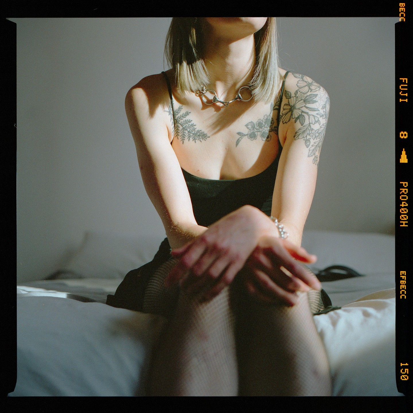 A woman with tattoos and a necklace sits on a bed in a sunlit room.