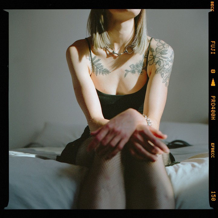 A woman with tattoos and a necklace sits on a bed in a sunlit room.