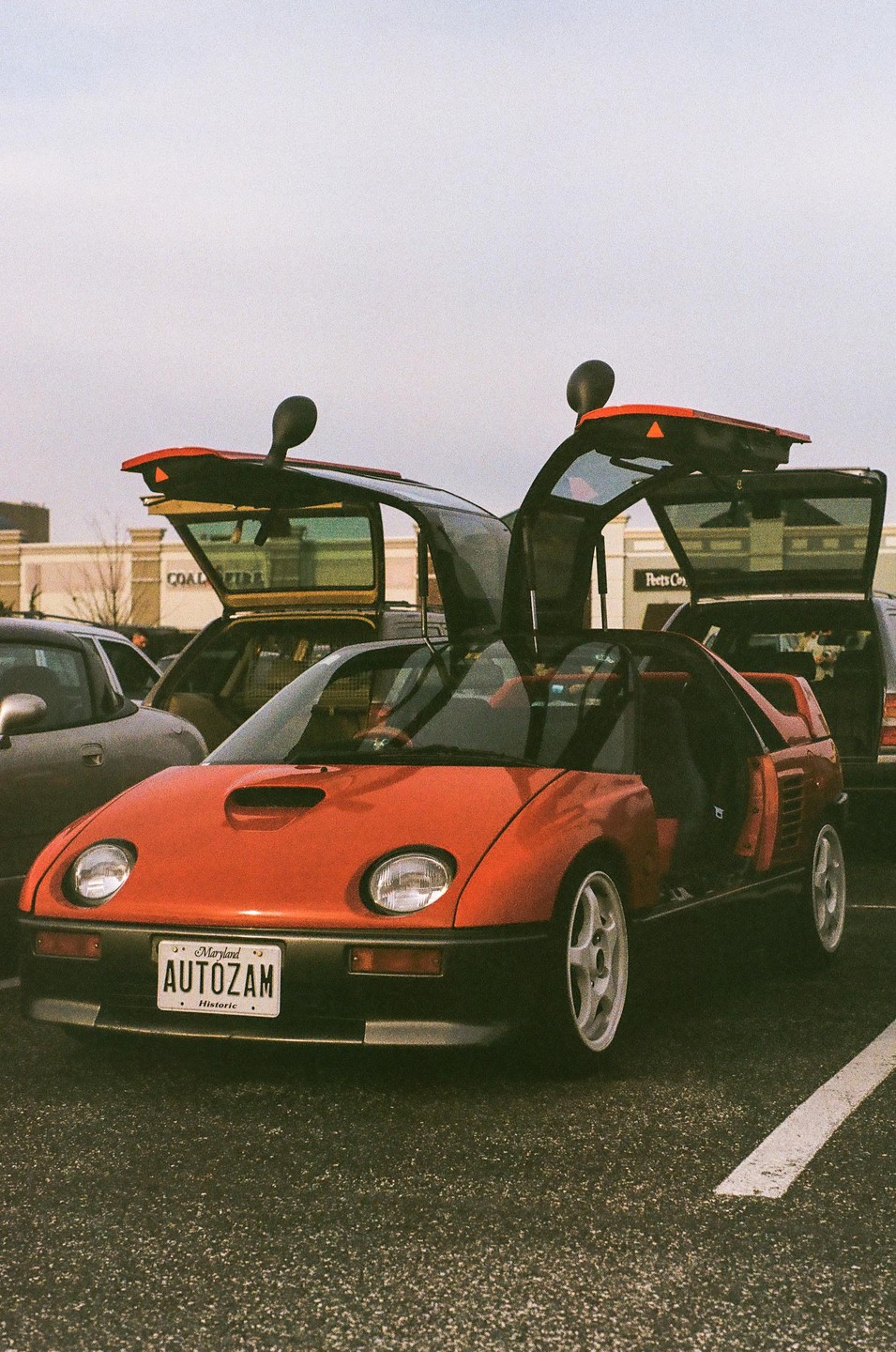 A red Autozam AZ-1 with its gull-wing doors open is parked in a lot.