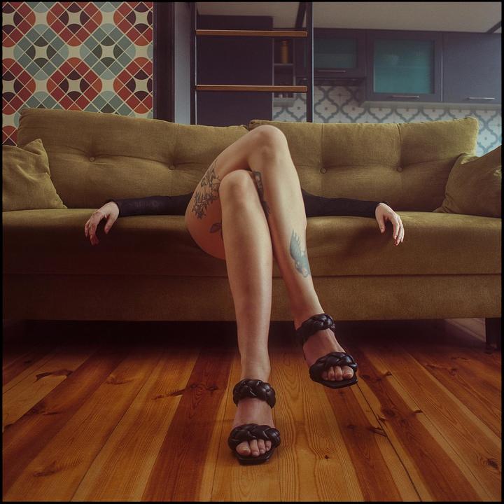 A woman with tattooed legs and black sandals is lying on a green sofa with her arms outstretched.
