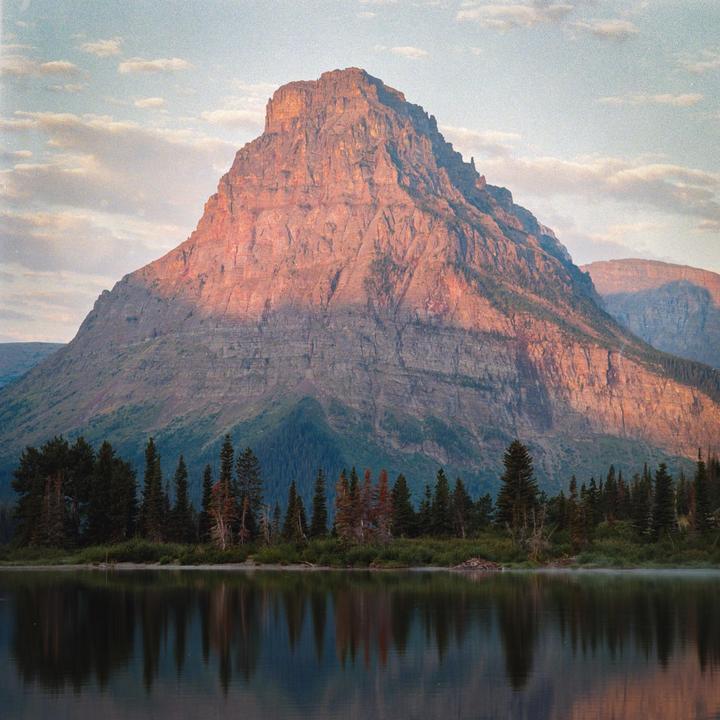 A large mountain with a forested base is reflected in a lake under a cloudy sky.