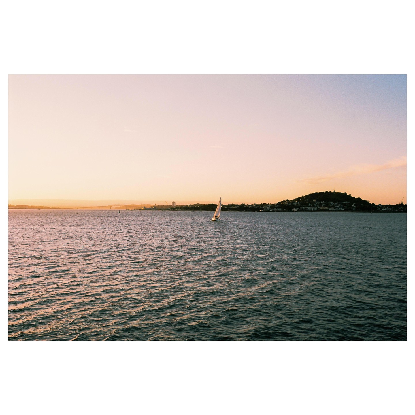 A sailboat glides across the water with a city and hills in the background at sunset.