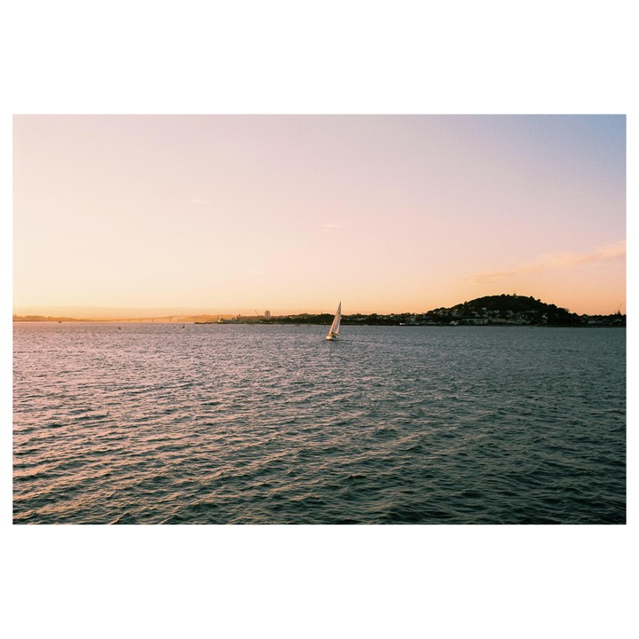 A sailboat glides across the water with a city and hills in the background at sunset.