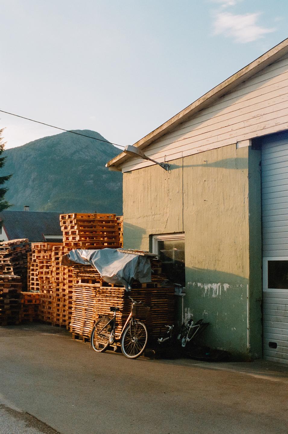 A bicycle is parked next to a building with stacks of wooden pallets and a mountain in the background.