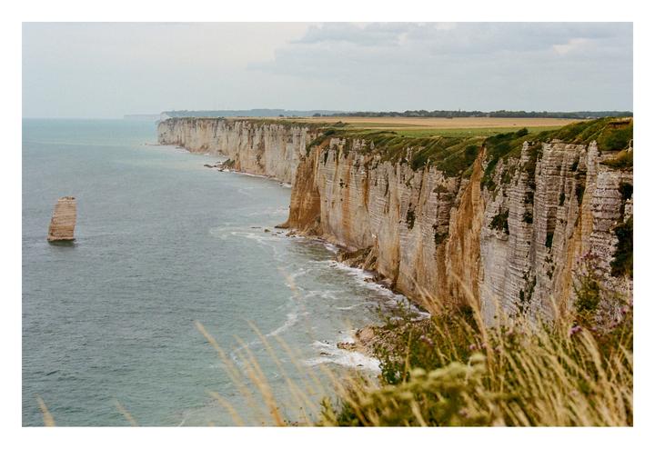 A long line of white cliffs with green tops stretches along the sea, with a single sea stack standing in the water.