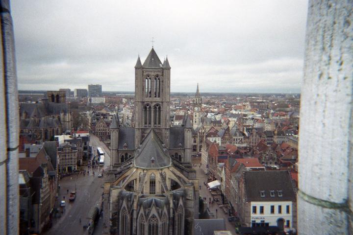 A large gothic cathedral with a tall bell tower stands in the center of a city with many buildings and a street with vehicles and pedestrians.
