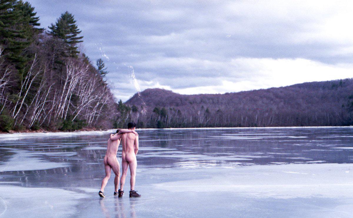 Two men stand naked on a frozen lake with a forest and mountain in the background.