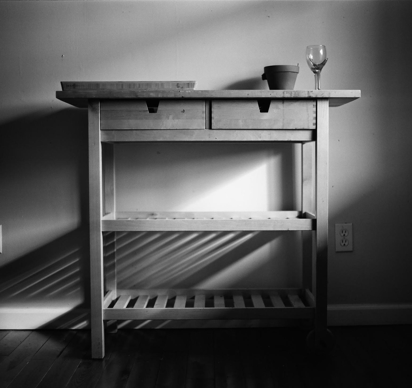 A wooden table with two drawers, a cutting board, a small pot, and a wine glass is illuminated by evening sun.