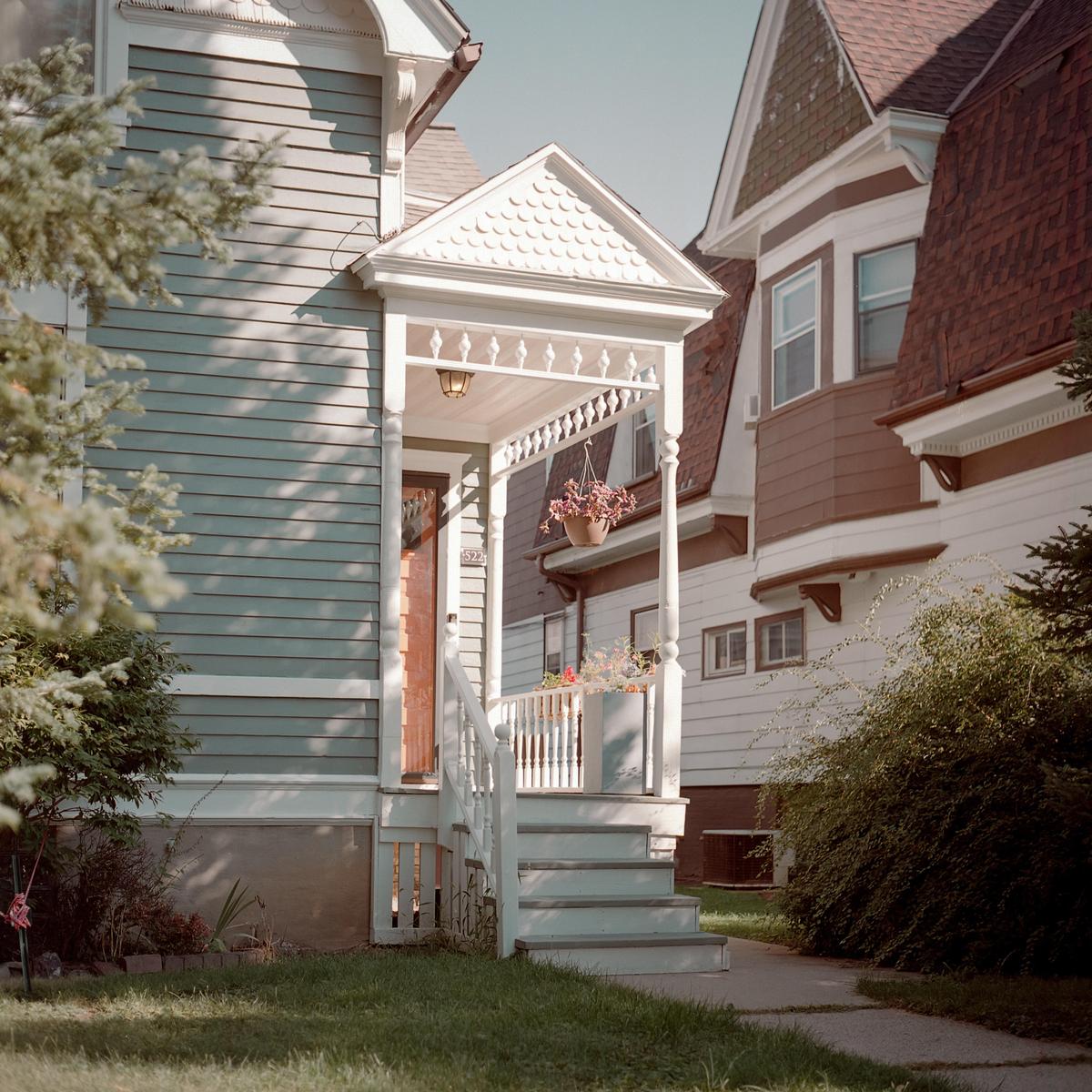 A white porch with steps leads to a house with light blue siding, next to a house with white siding and a brown roof.