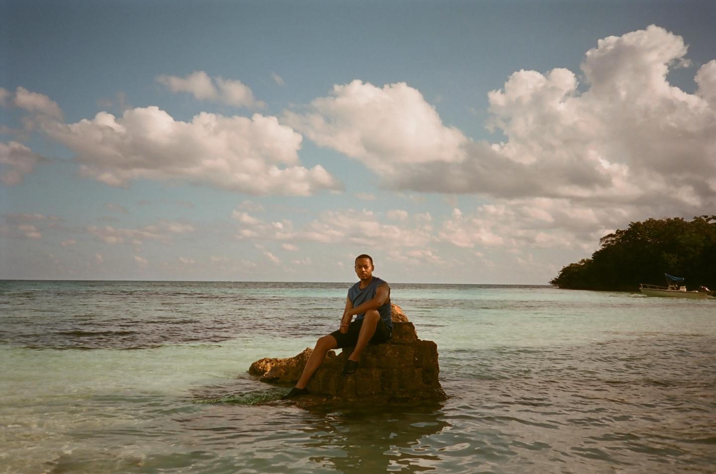 A man sits on a rock in the clear blue ocean with an island and boat in the background.