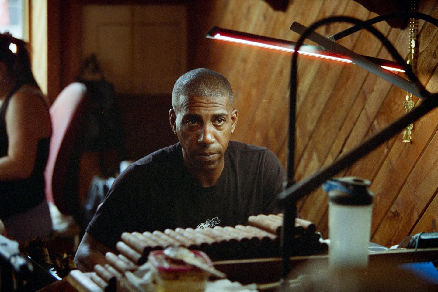 A man sits at a table with many cigars in front of him, with a woman in the background.