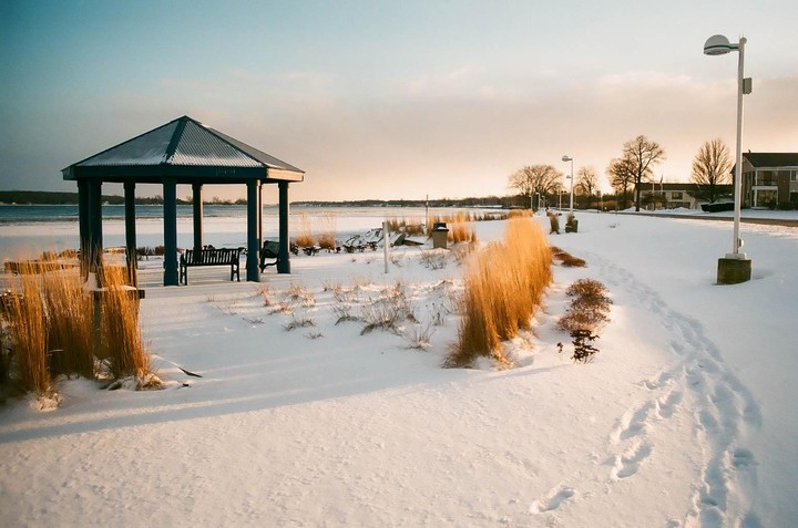 A snowy landscape features a gazebo by a body of water with a path leading away from it, illuminated by the warm light of sunset.