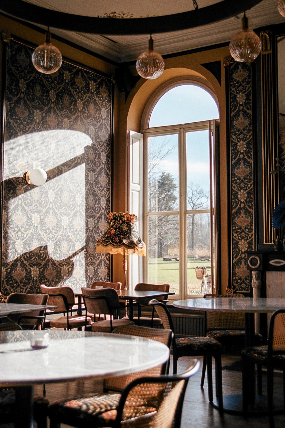 Sunlight streams through a tall arched window into a room with patterned wallpaper, round tables, and chairs.