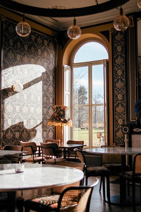 Sunlight streams through a tall arched window into a room with patterned wallpaper, round tables, and chairs.