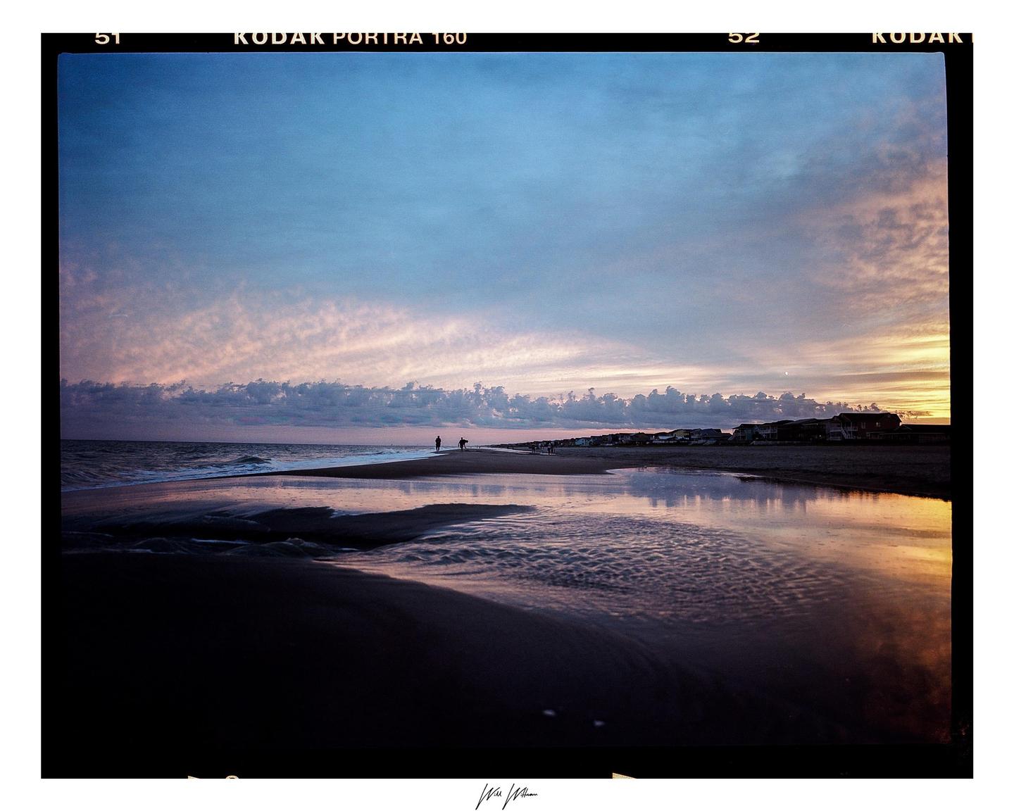 Two people walk along a beach at sunset, with houses visible in the distance.