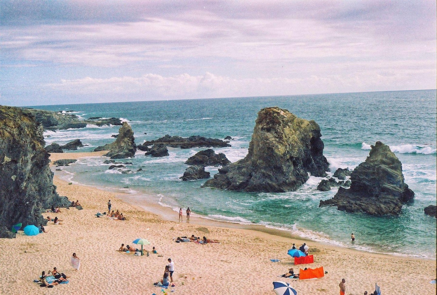 A sandy beach with many people and umbrellas is next to a rocky ocean with large rock formations.