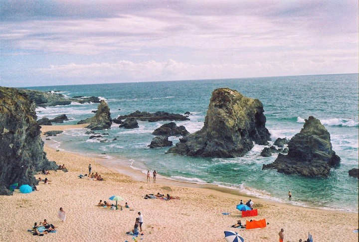 A sandy beach with many people and umbrellas is next to a rocky ocean with large rock formations.