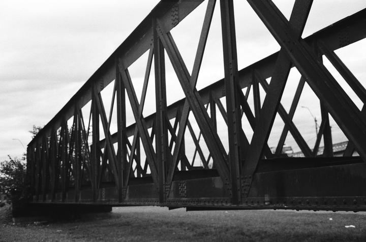 A black and white photograph shows the side of a metal truss bridge with a cloudy sky above.