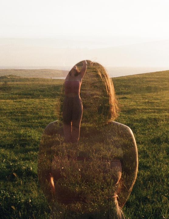 A double exposure shows a woman from behind superimposed with a woman with her arms raised in a grassy field at sunset.
