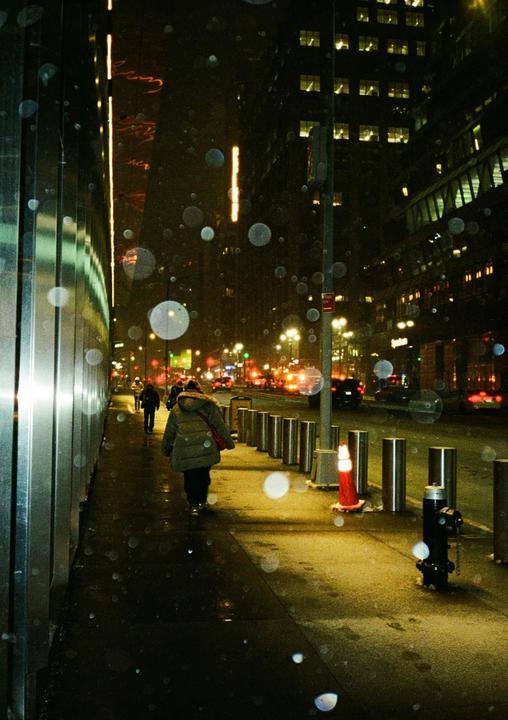 A person walks along a city sidewalk at night, with bright lights and reflections visible on the wet ground.