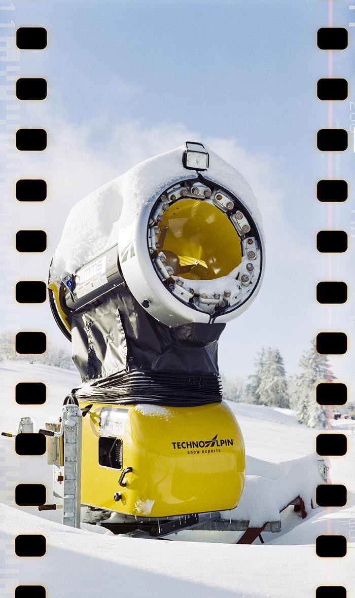 A yellow and white snow cannon stands in a snowy landscape with trees in the background.