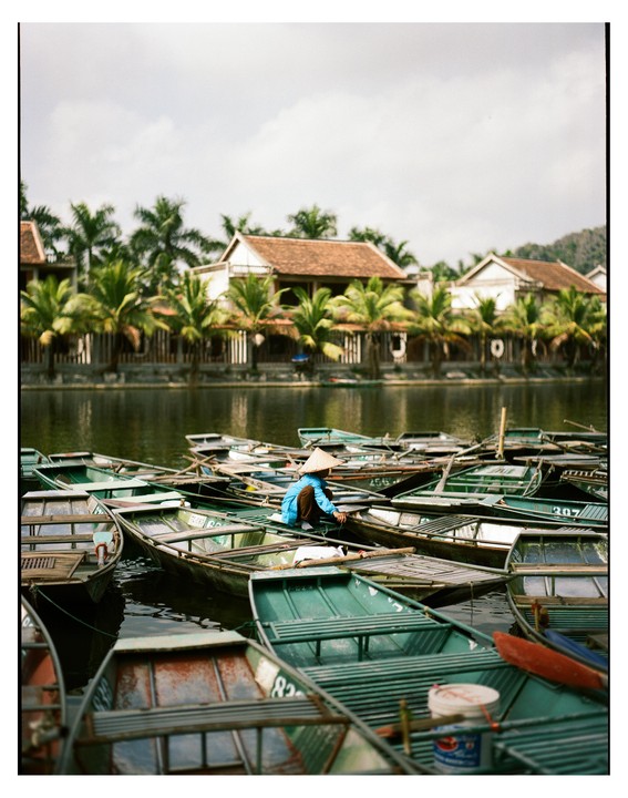 A person wearing a conical hat sits in a boat among many other boats on a body of water, with buildings and palm trees on the far bank.