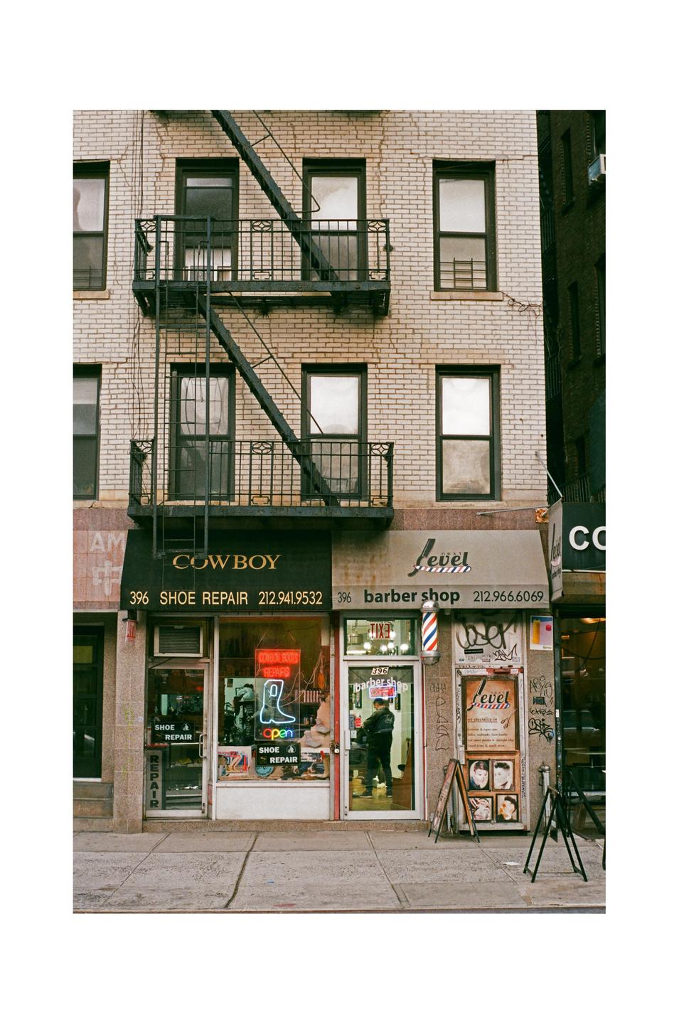 A brick building with a fire escape has a shoe repair shop and a barber shop on the ground floor.