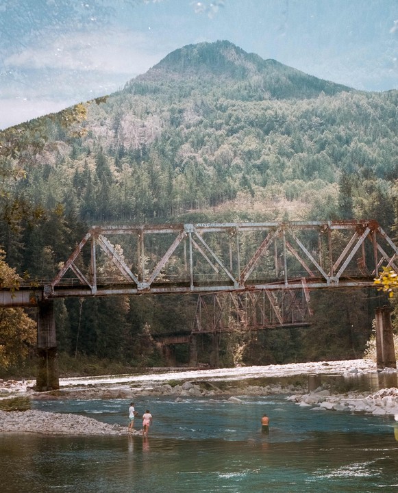 A group of people wade in a river beneath a metal bridge with a forested mountain in the background.