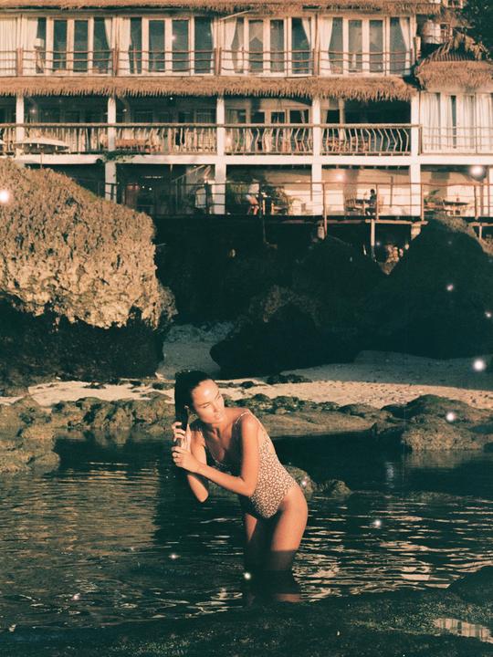 A woman in a swimsuit stands in the ocean with a multi-story building and rocks in the background.