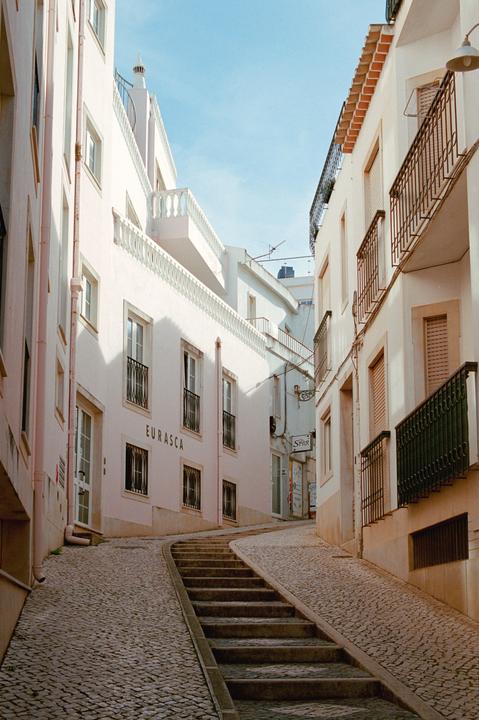 A narrow cobblestone street with a long set of stairs in the middle is lined with white buildings with balconies.
