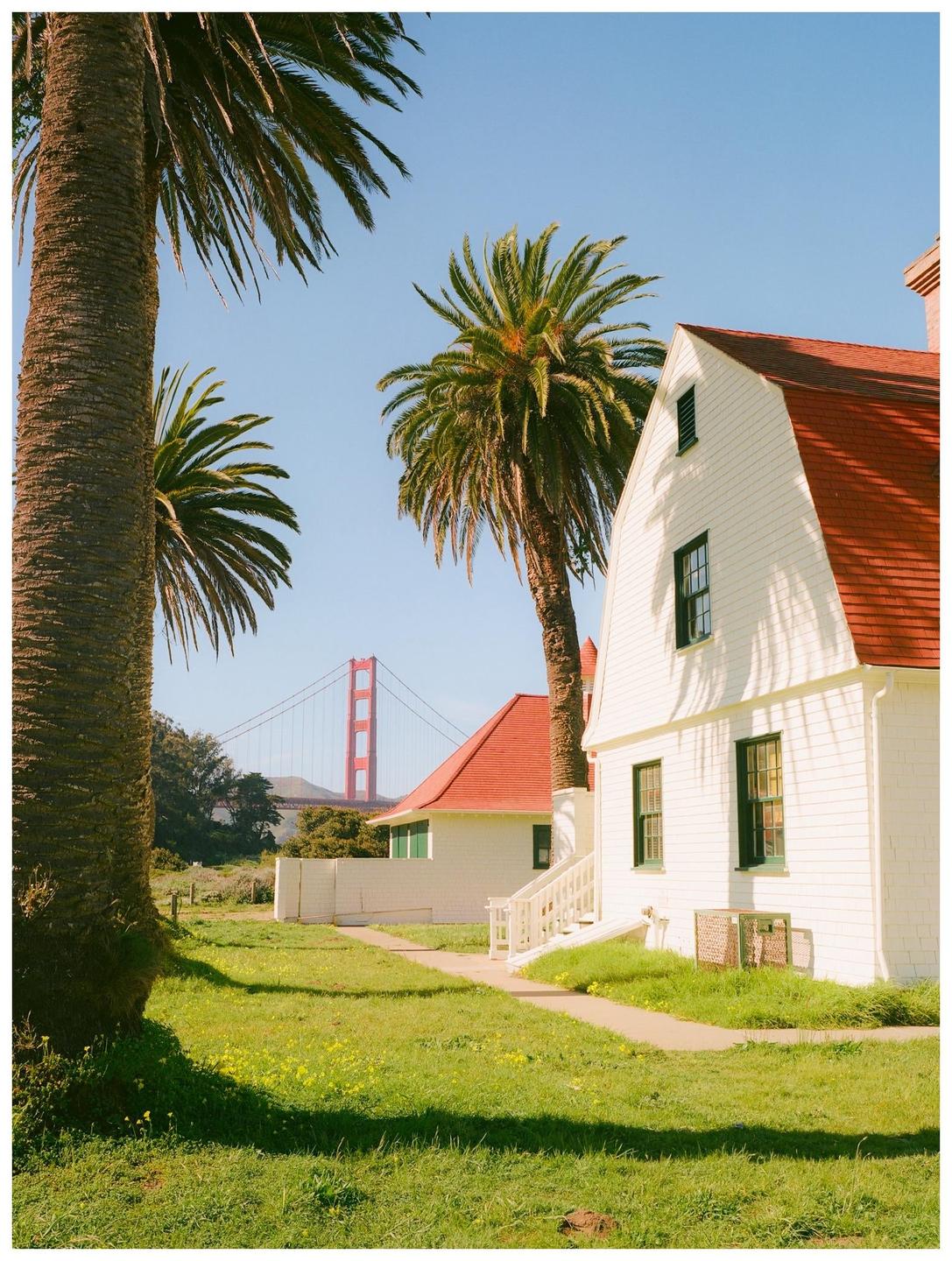 A white house with a red roof stands next to palm trees, with the Golden Gate Bridge visible in the distance.
