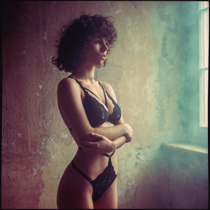 A woman with curly hair stands in black lingerie with her arms crossed, looking out a window.